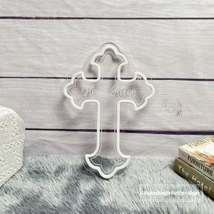 Puede incluir: Una cruz blanca iluminada con la inscripci&oacute;n "He is Risen". La cruz tiene un borde decorativo festoneado y se encuentra sobre un fondo claro de paneles de madera gris. La cruz est&aacute; sobre una superficie gris y esponjosa.