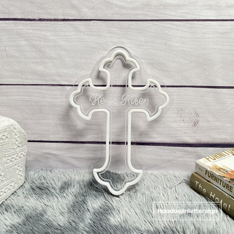 Puede incluir: Una cruz blanca iluminada con la inscripci&oacute;n "He is Risen". La cruz tiene un borde decorativo festoneado y se encuentra sobre un fondo claro de paneles de madera gris. La cruz est&aacute; sobre una superficie gris y esponjosa.
