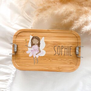 Könnte beinhalten: Eine rechteckige Bambus-Lunchbox mit Metallverschlüssen. Der Deckel zeigt eine Cartoon-Fee in einem rosa Kleid, die einen Zauberstab hält, mit dem Namen "SOPHIE" in Schwarz aufgedruckt. Die Lunchbox ist aus hellbraunem Bambus gefertigt.