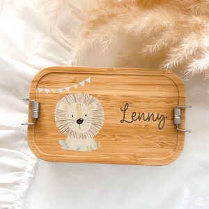 Könnte beinhalten: Eine rechteckige Bambus-Lunchbox mit einer Cartoon-Löwen-Illustration und dem Namen "Lenny" in Schreibschrift. Der Löwe ist beige mit einer hellbraunen Mähne. Die Lunchbox hat silberne Metallverschlüsse und ein kleines Banner.