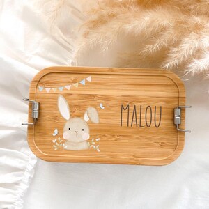 Könnte beinhalten: Eine rechteckige Bambus-Lunchbox mit einer Hasenillustration und dem Namen "MALOU" in Schwarz. Der Deckel zeigt einen Cartoon-Hasen, kleine Schmetterlinge und ein Banner mit Pastellflaggen. Metallverschlüsse sichern die Box.