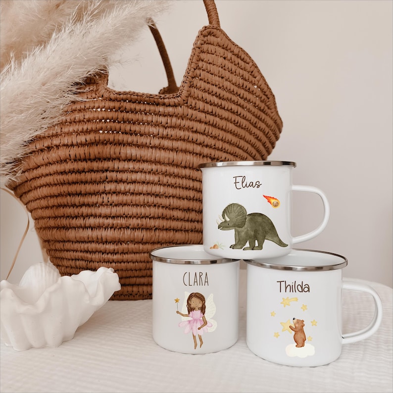 May include: Three white enamel mugs with silver rims, each featuring a unique illustration and name. One mug shows a green triceratops and the name Elias, another a fairy and the name Clara, and the third a brown bear and the name Thilda.