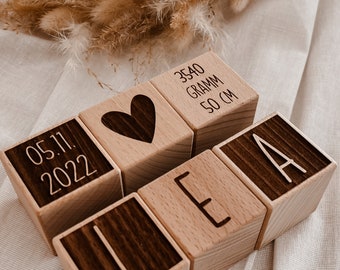 May include: Five light-colored wooden blocks arranged in two rows. The top row features a block with the date 05.11.2022, a block with a dark heart icon, and a block with the text 3540 GRAMM 50 CM. The bottom row features three blocks spelling LEA.