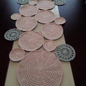 May include: A set of crocheted table mats and coasters in pink and blue. The mats are round and have a concentric circle design. The coasters are smaller and have a floral design.