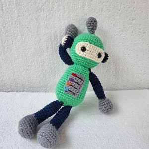 May include: A green crocheted robot toy with blue and gray legs and a white face with black eyes.