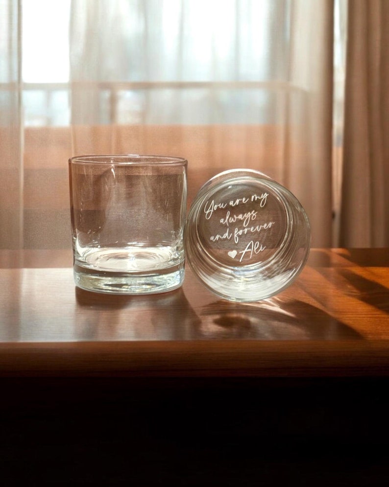May include: Two clear, lowball glasses on a wooden surface. One glass is upright, the other is lying on its side, revealing the etched text "You are my always and forever Ali" with a heart symbol. The background features a sheer curtain.