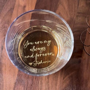 May include: A clear glass tumbler with the inscription "You are my always and forever Stephanie" in a gold script. The glass is filled with a light-colored liquid and sits on a dark wooden surface.