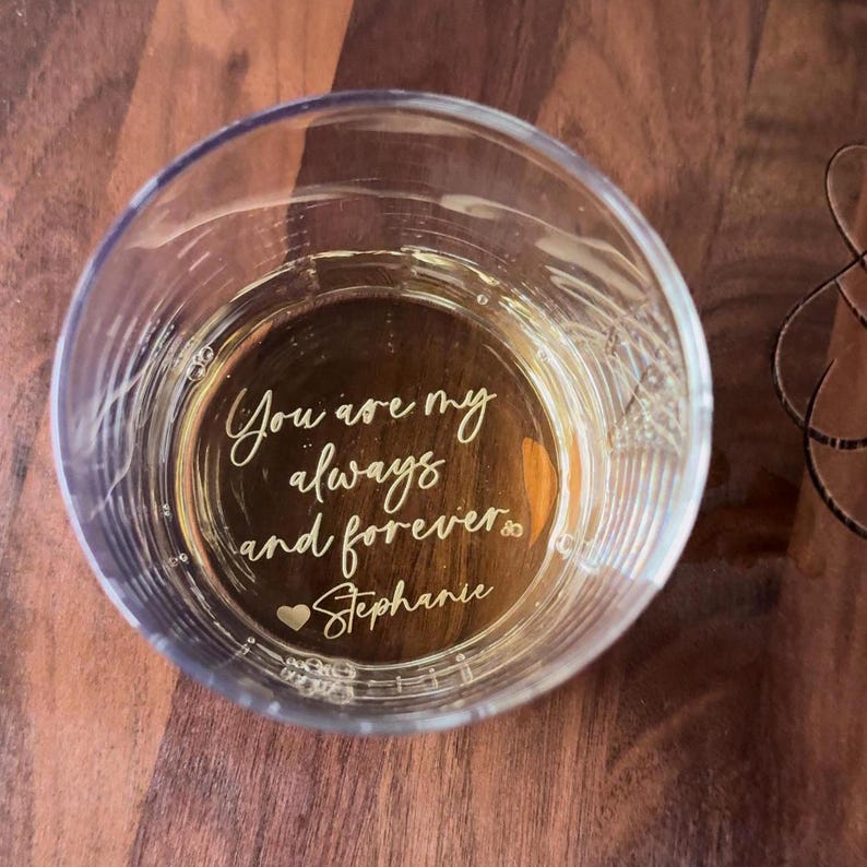 May include: A clear glass tumbler with the inscription "You are my always and forever Stephanie" in a gold script. The glass is filled with a light-colored liquid and sits on a dark wooden surface.