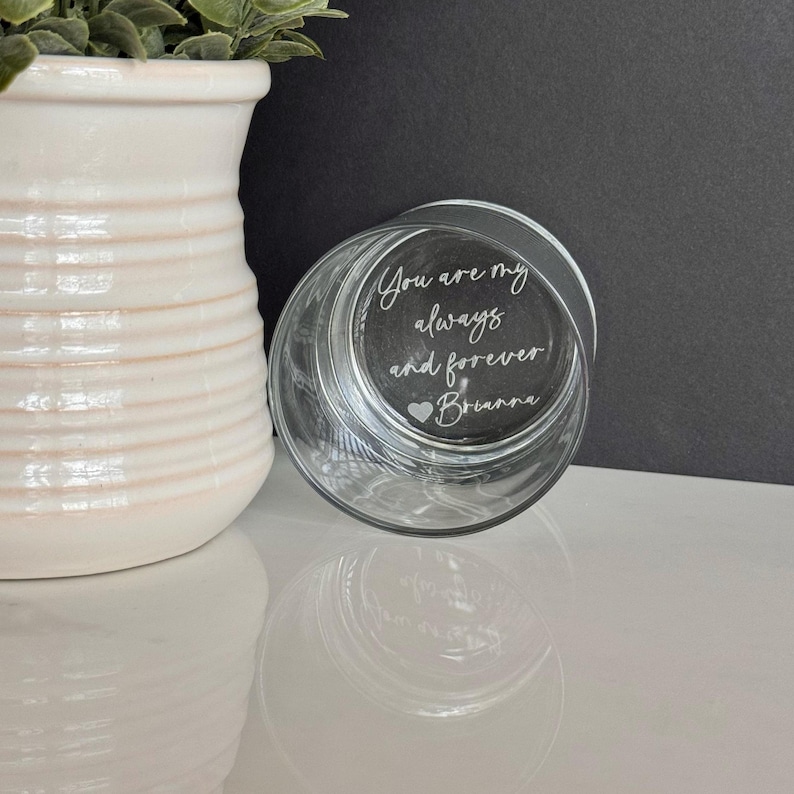 May include: A clear glass tumbler with the words "You are my always and forever Brianna" etched on the bottom. The glass is partially visible, with a white vase and greenery in the background.