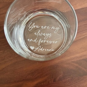 May include: A clear glass tumbler with the etched inscription "You are my always and forever Adriana" in a cursive font. A small heart symbol is included. The glass is set on a brown wooden surface.