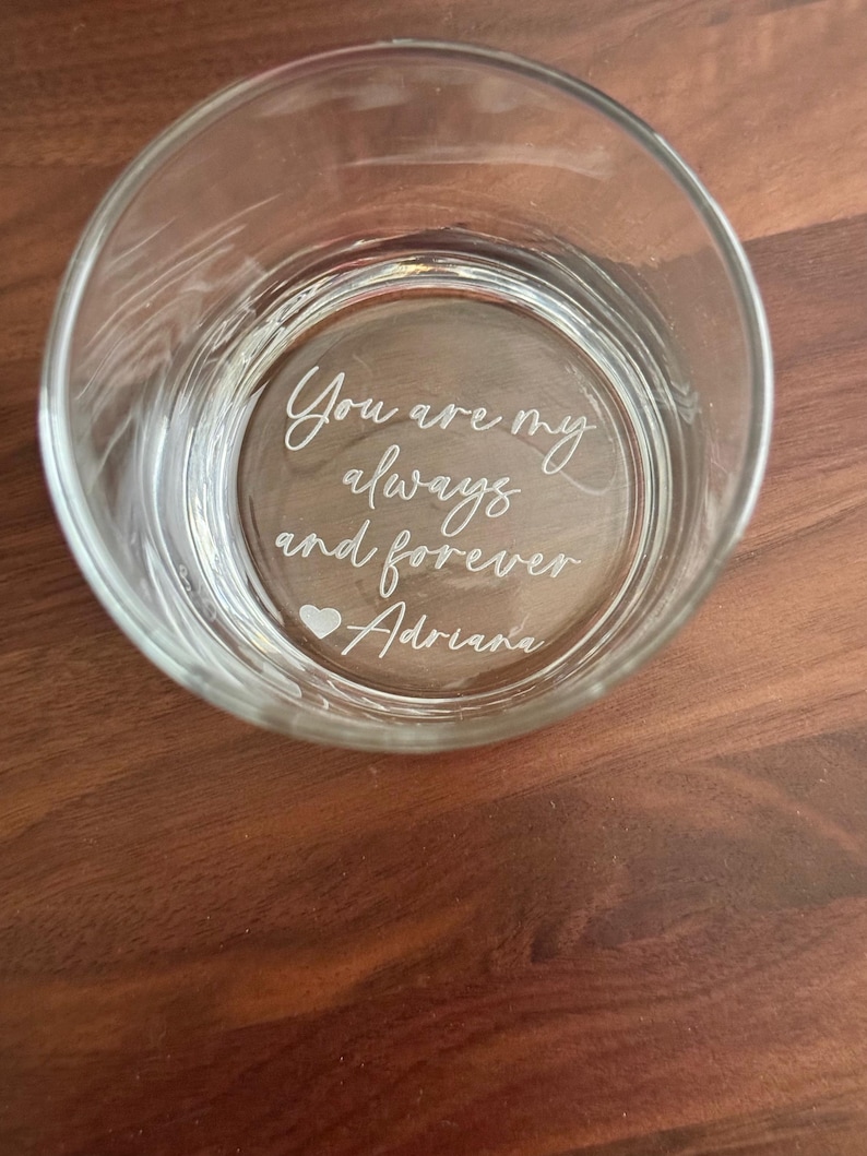 May include: A clear glass tumbler with the etched inscription "You are my always and forever Adriana" in a cursive font. A small heart symbol is included. The glass is set on a brown wooden surface.