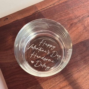 May include: A clear glass votive candle holder with the words "Happy Valentine's Day Handsome Delmy" etched on the bottom. The candle holder sits on a dark brown wooden surface.
