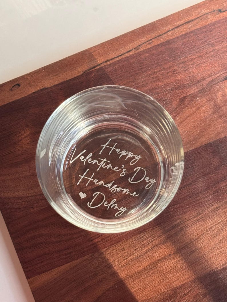 May include: A clear glass votive candle holder with the words "Happy Valentine's Day Handsome Delmy" etched on the bottom. The candle holder sits on a dark brown wooden surface.