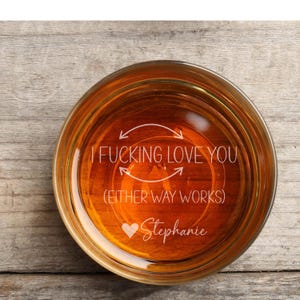 May include: A round glass filled with a golden liquid, likely a beverage, with etched text. The text reads "I FUCKING LOVE YOU (EITHER WAY WORKS)" with a heart and the name "Stephanie" below. The glass sits on a wooden surface.