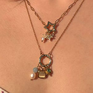 May include: A layered gold-tone necklace set featuring two chains. The shorter chain has a star-shaped pendant with small clear crystals. The longer chain features a circular ring pendant adorned with a white pearl, a grey bead, a square tan charm, and a small green stone.