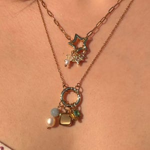 May include: A layered gold-tone necklace set featuring two distinct chains. The shorter chain has a star-shaped pendant with small clear stones. The longer chain features a circular ring charm adorned with a white pearl, a grey bead, a square tan charm, and a small green stone.