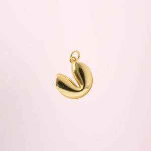 Puede incluir: Un dije dorado con forma de galleta de la fortuna, con un lazo para un collar o pulsera. El dije es liso y brillante, con la forma clásica de una galleta de la fortuna. El fondo es rosa pálido.