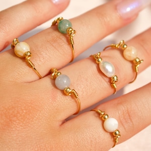 Op de afbeelding: Een hand toont verschillende gouden draadringen, elk versierd met een andere edelsteen of parel. De ringen hebben kralen in verschillende kleuren, waaronder groen, wit en perzik, en een enkele parel. De ringen zijn delicaat en elegant.