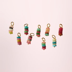May include: A selection of colourful gemstone charms with gold-coloured loops. Each charm features stacked, round, polished stones in various colours, including pink, green, blue, and yellow. The charms are set against a soft pink background.