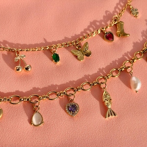 May include: Two gold-toned charm bracelets featuring various pendants including a butterfly, a cowboy boot, cherries, a fish, a heart, and several colourful gemstones. The chains are constructed with linked metal loops and secure clasps.