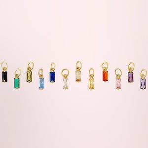 Op de afbeelding: Een collectie van twaalf kleine goudkleurige bedels, elk voorzien van een rechthoekige baguette-geslepen edelsteen in een andere kleur, waaronder zwart, groen, olijf, lichtblauw, donkerblauw, transparant, amber, geel, oranje, roze, paars en violet.