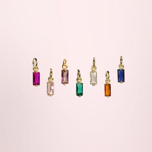 Op de afbeelding: Zeven goudkleurige rechthoekige bedels met kleurrijke, edelsteenachtige stenen in tinten roze, paars, groen, helder, oranje en blauw. Elke bedel heeft een kleine lus voor bevestiging. De bedels worden getoond tegen een zachtroze achtergrond.