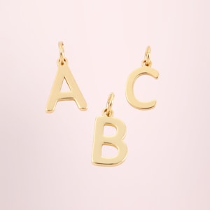 May include: Three gold-colored letter charms: A, B, and C. Each letter has a small loop at the top for attaching to a chain or bracelet. The letters are displayed against a soft pink background.