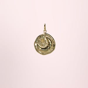 May include: A gold-colored pendant featuring a crescent moon and a star design. The pendant has a textured surface and a small loop at the top for a chain. The background is a soft pink.