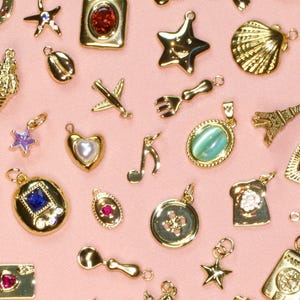 May include: A collection of gold-toned metal charms in various shapes including stars, hearts, a seashell, an aeroplane, a fork, a musical note, a dice, a passport, and an Eiffel Tower, some featuring embedded colourful gemstones and pearls.