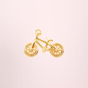 Peut inclure: Un charm de vélo doré avec des rayons et un cadre détaillés. Le charm est une représentation miniature d'un vélo, idéal pour les bijoux ou les projets d'artisanat. Le charm de vélo est posé sur un fond rose clair.