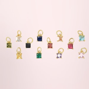 May include: A collection of twelve small square-cut gemstone charms, each set in a gold-toned metal frame with a circular jump ring attached at the top. The gemstones feature a variety of colors including clear, black, blue, green, red, and pink.