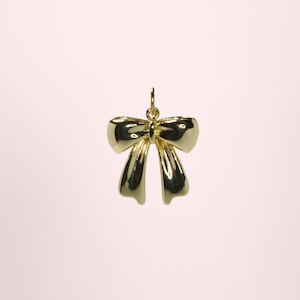 Peut inclure: Un pendentif en forme de nœud doré avec une boucle en haut pour une chaîne. Le nœud a une finition brillante et est suspendu sur un fond rose clair. Le pendentif est un bijou.