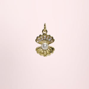 May include: A gold-coloured seashell pendant with a pearl and sparkling accents. The pendant features an open clam shell design with a central pearl and small, clear crystals. The jewellery is set against a soft pink background.