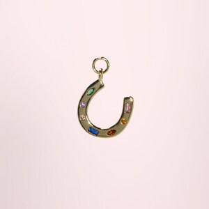 May include: A gold-colored horseshoe pendant with a loop at the top for hanging. The horseshoe is adorned with colorful gemstones in various shapes and sizes, including blue, pink, and orange. The pendant is set against a soft pink background.
