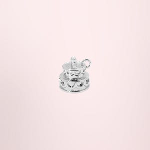 Puede incluir: Un pequeño dije de metal plateado brillante con forma de pastel de dos niveles. El dije presenta un diseño de borde festoneado alrededor de la base y el nivel superior, con una pequeña proyección vertical en la parte superior y una argolla.