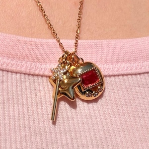 May include: A gold-tone necklace featuring a pendant cluster with a star-shaped charm adorned with clear crystals, a wand-shaped charm, and a circular charm containing a square red stone surrounded by small clear crystals on a fine chain.
