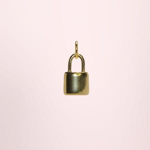May include: A small, gold-colored padlock pendant with a rounded rectangular body and a curved handle. The pendant hangs from a small loop. The background is a soft pink.