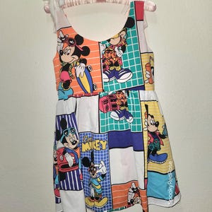 May include: A sleeveless dress featuring a patchwork design with various Mickey Mouse graphics. The dress showcases Mickey in different poses, set against colorful backgrounds of orange, blue, green, and white. The dress is hanging on a pink hanger.