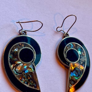 Op de afbeelding: Een paar zilveren oorbellen met een zwart en iriserend abalone-schelpontwerp. De oorbellen zijn spiraalvormig en hebben een haaksluiting.