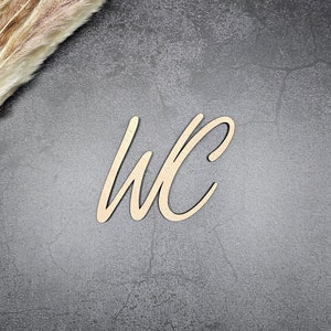 May include: A wooden sign with the letters "WC" in a cursive font, on a dark gray background with dried pampas grass in the top left corner.