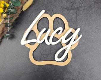 May include: A wooden paw print with the name "Lucy" in white script. The paw print is light brown, and the name is layered on top. The background is a dark gray, textured surface.