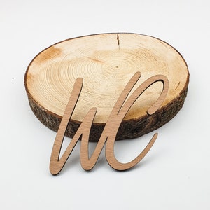 May include: Wooden cutout of the letters "WC" in a cursive font, sitting on a round wooden slice.