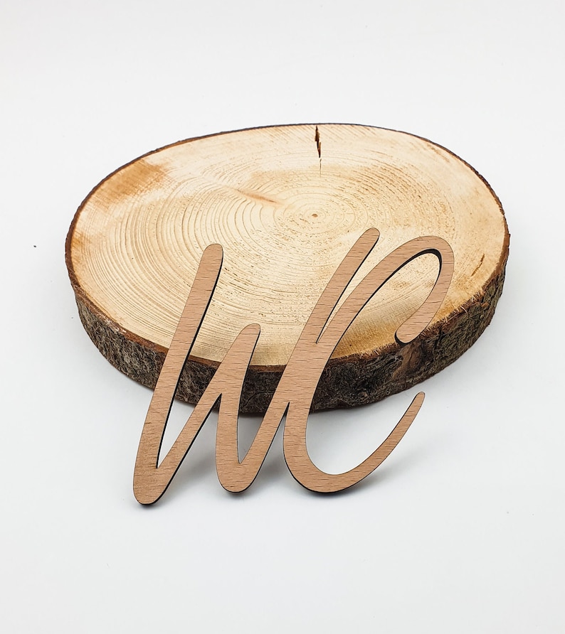 May include: Wooden cutout of the letters "WC" in a cursive font, sitting on a round wooden slice.