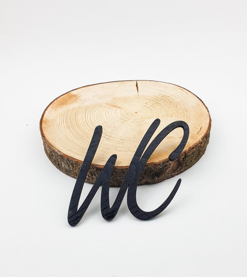 May include: Black wooden cutout of the letters "WC" on a natural wood slice.