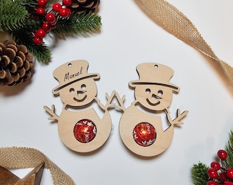 May include: Two light-colored wooden snowman ornaments with top hats, each featuring a circular cutout holding a red spherical chocolate. One snowman is engraved with the name Manuel. Both figures have simple stick arms and a smiling face design.