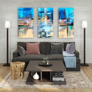 Op de afbeelding: Een donkergrijze bank met twee roze en twee grijze sierkussens, een lichtblauwe plaid, een vierkante salontafel van donker hout met een witte vaas en drie verticale abstracte schilderijen in blauw, goud en wit aan een bakstenen muur.