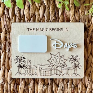 May include: A rectangular light-colored wooden countdown sign featuring the laser-engraved text THE MAGIC BEGINS IN and POP CENTURY. It includes a white rectangular dry-erase area, the word Days in a stylized font, and palm tree illustrations.