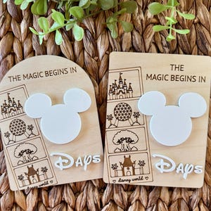 May include: Two light-colored wooden countdown signs featuring the text THE MAGIC BEGINS IN and DAYS. Each sign displays laser-engraved outlines of four theme park landmarks and a white acrylic Mickey Mouse head silhouette in the center.