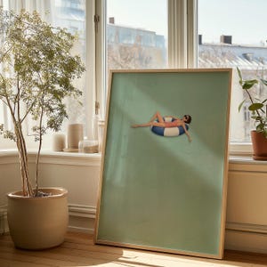 Pu&ograve; includere: Una stampa artistica da parete rettangolare incorniciata con sfondo verde salvia e un'illustrazione centrale di una figura in costume da bagno rosa e occhiali da sole, sdraiata su un salvagente circolare a righe blu e bianche.