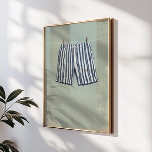 May include: Framed art print featuring a pair of blue and white striped shorts hanging on a clothesline. The shorts are the focal point, set against a muted green background. The artwork is framed in a gold-colored frame.